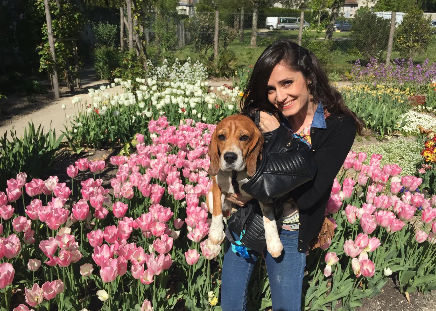 Marina posando para la foto, cogiendo en brazos a su perro Milo, al fondo de la imagen se muestran muchos tulipanes de diferentes colores y más plantas.